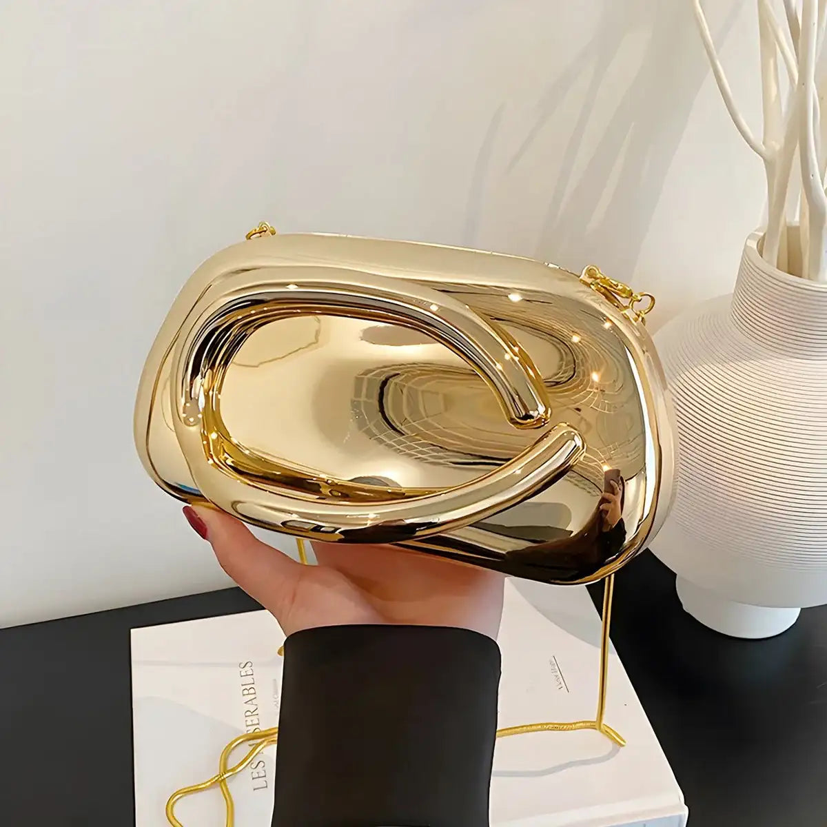 Gold clutch held by a hand on a white surface with a lamp and vase in the background